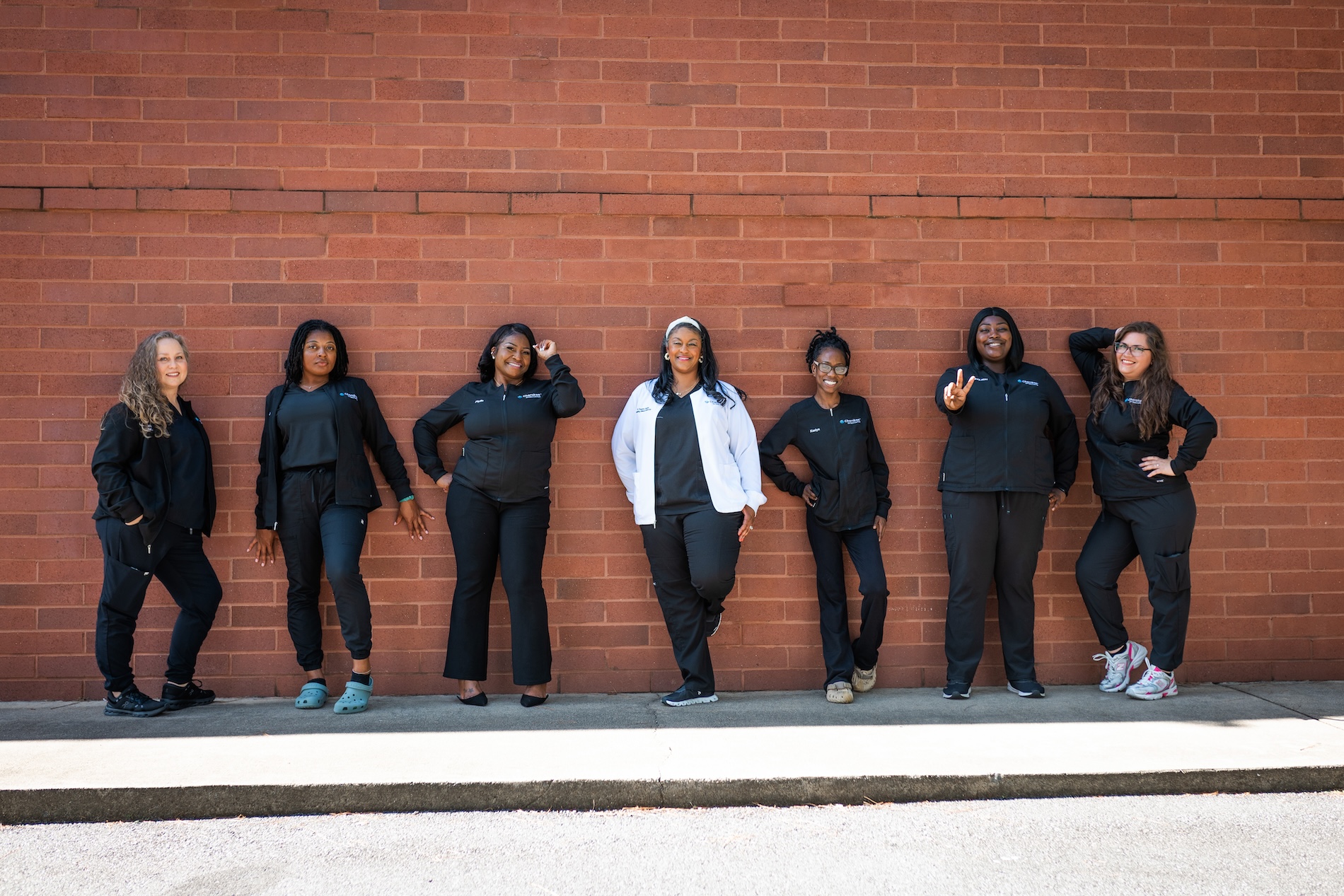 The image shows a group of seven individuals posing together with a brick wall behind them, wearing matching black jackets with visible name tags, and smiling at the camera.