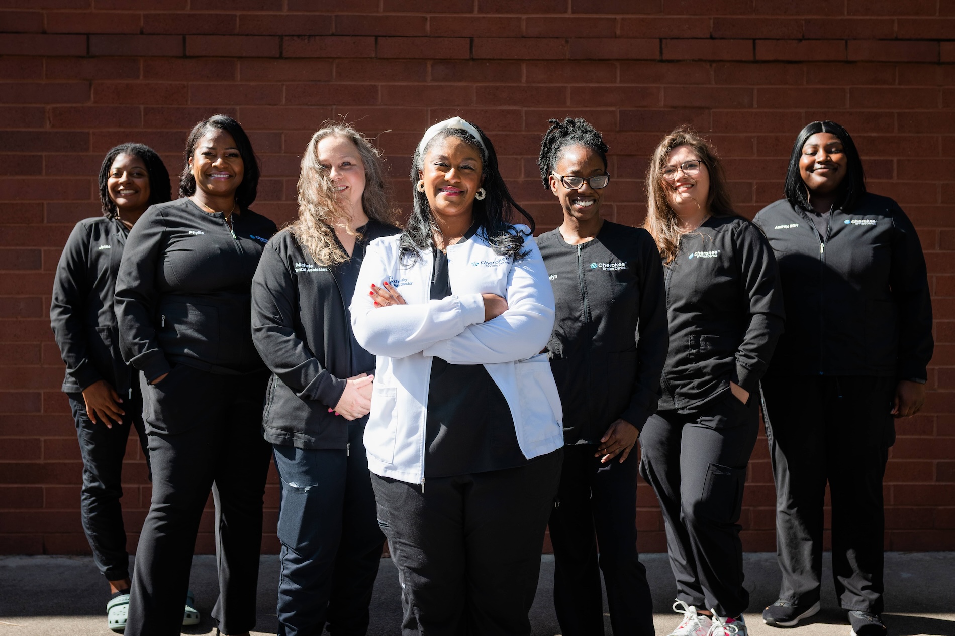 A group of women wearing scrubs pose together outdoors, with one woman standing slightly apart from the others.