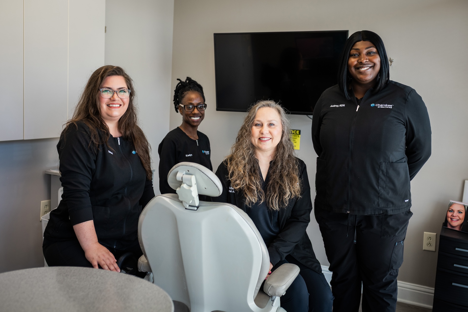 The image shows four individuals posing with a dental chair in an office setting, with a sign indicating  Dental Hygiene.