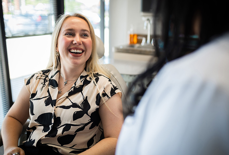 A woman with blonde hair wearing a black and white top, seated at a dental office chair, smiling and looking towards the camera, while another person is partially visible on the right side of the frame.