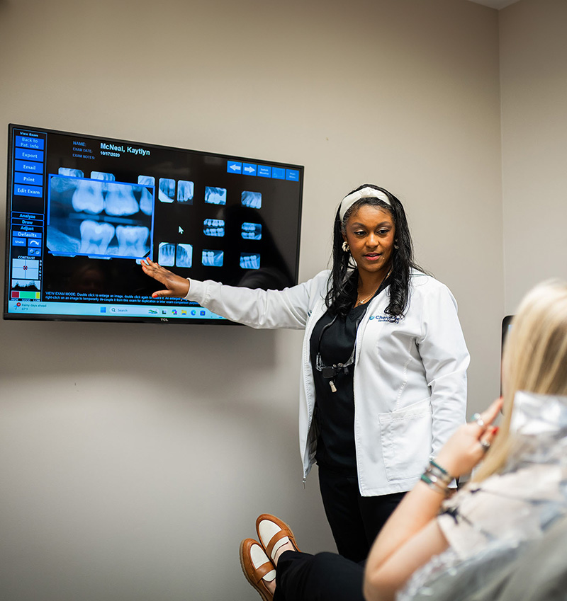 A dental professional standing next to an X-ray monitor with a patient s mouth open on the screen, pointing at it.