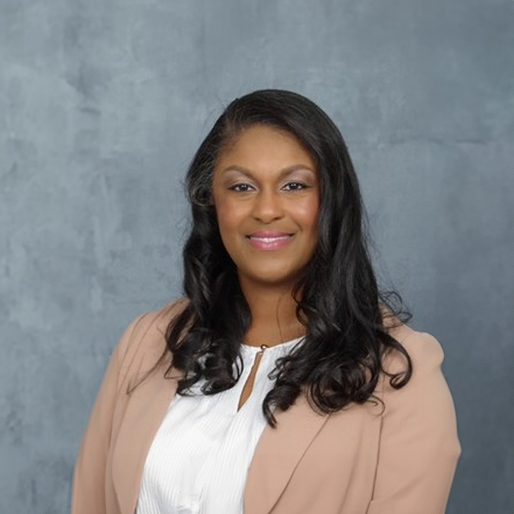 The image shows a woman posing for a portrait with a neutral background. She has long hair, is wearing a light-colored blazer over a white top, and has a warm smile on her face.