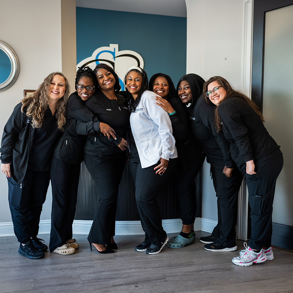 Group of smiling women posing together in professional attire, likely at a dental office, with some holding phones and others hugging each other.