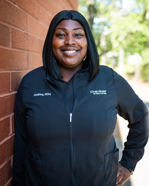 A smiling woman wearing a black jacket with a name tag, standing against a brick wall.