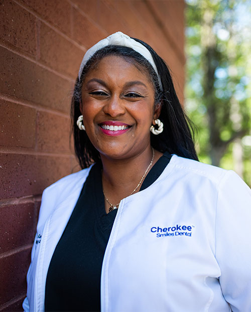 The image shows a woman wearing a white lab coat with a name tag, smiling at the camera. She has dark hair, is wearing glasses, and has a headband. In the background, there s a brick wall and trees can be seen.
