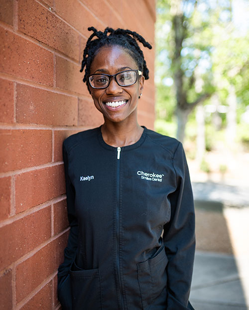 Woman wearing glasses and smiling at camera, leaning against brick wall with black jacket and name tag visible.