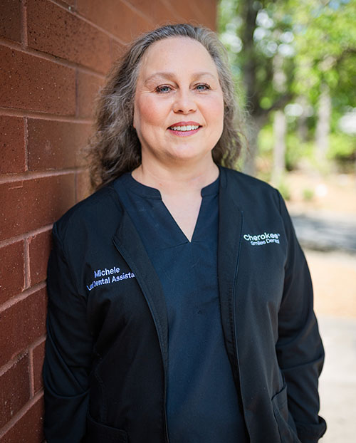 The image features a woman standing against a brick wall, wearing a black coat with a name tag on her left side, smiling at the camera.