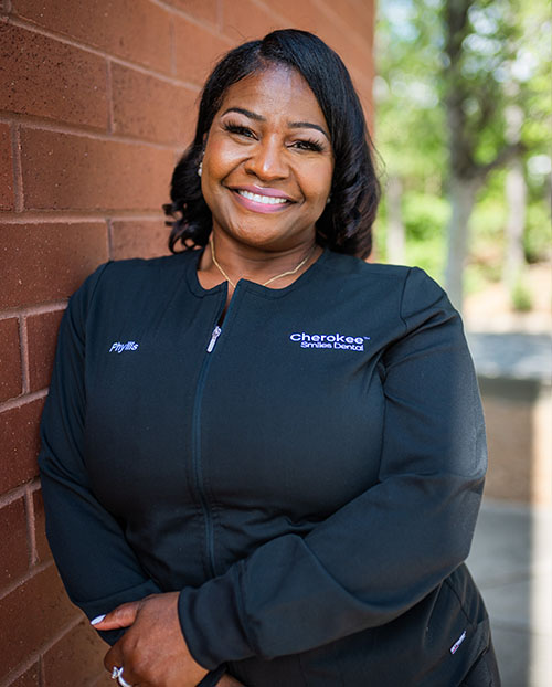 The image features a woman standing against a brick wall with a smile on her face, wearing a black jacket with a logo on the left chest area and a name tag that reads  Cherokee.