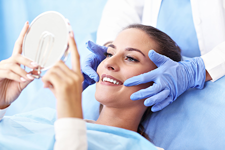 A woman lying on a dental chair with a magnifying mirror held over her face by a dental professional, both wearing blue gloves and masks.