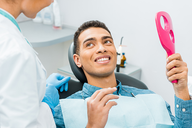 The image depicts a dental professional with a patient sitting in a chair, both wearing face masks, in a dental office setting with a red light on a device near the patient s mouth.
