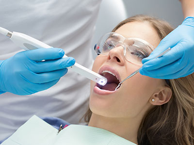 An adult female patient receiving dental care with a dental device inserted into her mouth, attended by a dental professional using an electronic tool, all set against a clinical background.